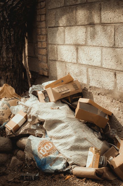 A pile of discarded cardboard boxes, some flattened and others partially intact, along with plastic packaging and a large woven sack, all resting on uneven ground next to a brick wall and a tree trunk